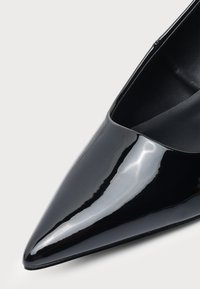 Gros plan d'une chaussure d'escarpin noire en cuir verni brillant, bout pointu, surface lisse et ouverture basse.