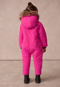 Rozā izolēts sniegapārslas kostīms ar mākslīgo kažokādu apdari kapucei, elastīgiem roku galiem un melnām zābakiem. Uz aizmugures ir zīmola nosaukums. Gluda, ūdensnecaurlaidīga materiāla.