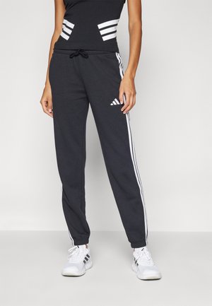 Person iført sorte Adidas træningsbukser med hvide sidelommer og logo, kombineret med hvide og sorte Adidas sneakers på en ensfarvet baggrund.