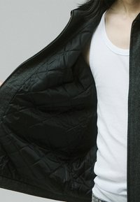 Chaqueta de arcade con exterior de lana, forro negro acolchado, cierre delantero con cremallera y sin hardware visible. El interior muestra una textura acolchada.