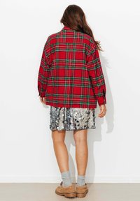 Chemise à carreaux rouges et verts à manches longues, associée à une jupe en sequins argentés. Le modèle porte des chaussettes grises et des chaussures beige clair. Le fond est blanc.