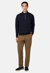 Jūras spēku zip-aizdare džemperis mīkstā audumā, apvienots ar brūnām biksēm; modelis valkā tumšus sporta apavus, stāvot ar vienu roku kabatā, vienkāršs fons.