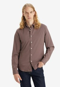 Brun langærmet skjorte med knapper og krave, der har hvide knapper og en glat tekstur, båret over mørke denim jeans.