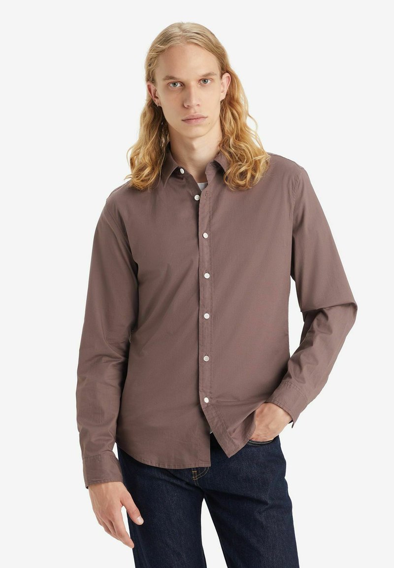 Brun langærmet skjorte med knapper og krave, der har hvide knapper og en glat tekstur, båret over mørke denim jeans.