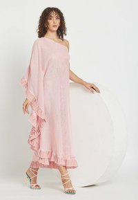 Mujer con cabello rizado junto a un gran círculo blanco, vestida con un vestido rosa de un solo hombro con bordes fruncidos y tacones altos pastel con tiras.