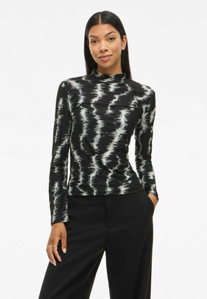 Femme aux longs cheveux noirs portant un haut à motifs noir et blanc à manches longues et un pantalon noir à larges jambes, se tenant avec une main dans la poche.