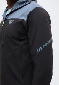 Κοντινό πλάνο ενός ατόμου που φοράει μαύρο και μπλε μπουφάν Dynafit με λογότυπο στο στήθος και όνομα μάρκας στο πάνω μέρος του μανικιού.