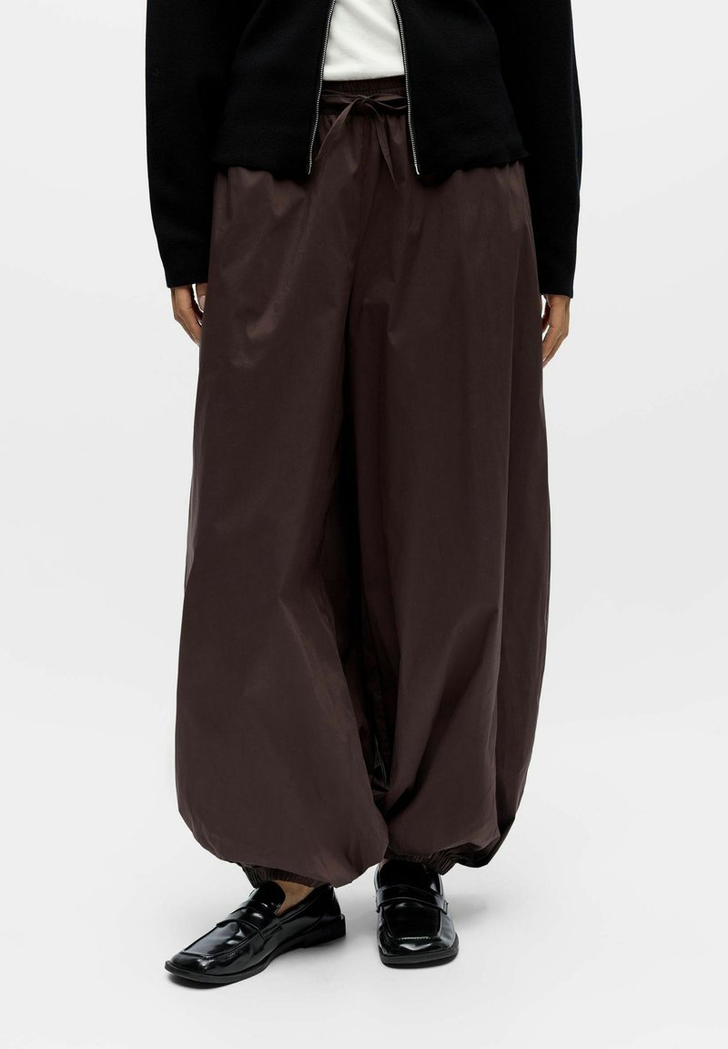 Pantalons marron amples en matière légère, dotés d'une taille élastique avec cordon de serrage et de poignets froncés, associés à des chaussures noires.