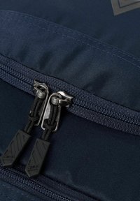 Gros plan sur un sac à dos en tissu navy avec des doubles zips, des tirettes noires et des accents texturés. Surface lisse avec des détails de couture visibles.