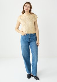 Beige gebreide mouwloze top met kabelpatroon en hoge halslijn, gecombineerd met hoge taille blauwe rechte jeans en zwarte schoenen.