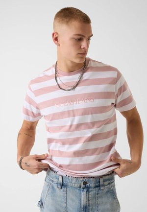 Giovane uomo con maglietta a righe rosa e bianche "Guess Jeans" e jeans azzurri, che sistema l'orlo della maglietta, guardando in basso verso sinistra.