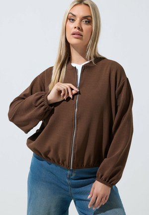 Lyshåret kvinde iført brun lynlåstrøje med elastiske manchetter, blå jeans, stående mod ensartet lyst baggrund, holder lynlåsen halvvejs oppe.