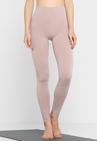 En person står barfota på en grå yogamatta iförd högmidjade, ljus mauve leggings i full längd mot en enfärgad vit bakgrund.