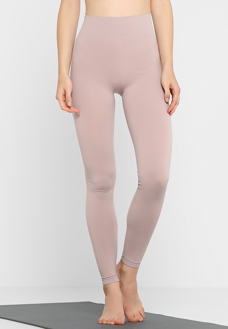 En person står barfota på en grå yogamatta iförd högmidjade, ljus mauve leggings i full längd mot en enfärgad vit bakgrund.