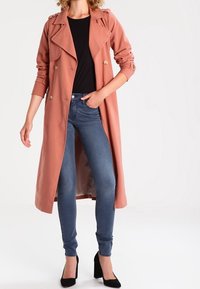 Gabardina de tejido coral suave, con diseño de doble botonadura, grandes solapas y botones en tono dorado. Combinada con jeans ajustados azules.