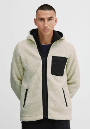 Fleece jacket in cream with a black zippered chest pocket, contrast black trims, and a hood. Textured material and side pockets.