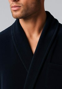 Primo piano di un uomo con barba curata che indossa una vestaglia blu navy scuro con colletto a scialle e taschino.