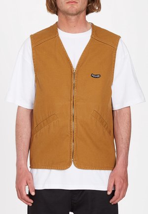 Homme portant un gilet marron zippé sur un T-shirt blanc à manches courtes, debout avec les bras détendus le long du corps.