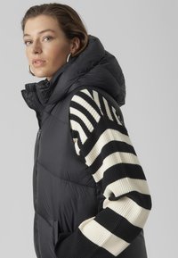 Veste sans manches noire avec un col montant et des manches en tricot rayées crème et noires. Le matériau est lisse avec une texture rembourrée.