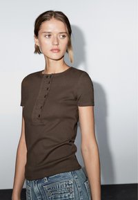 Top marrone a maniche corte con scollatura rotonda e pattina con bottoni. Il tessuto sembra a coste, abbinato a jeans in denim blu con tasche.