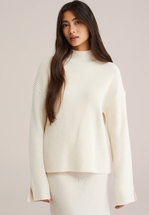 Jeune femme aux cheveux longs et foncés portant un pull en maille côtelée crème et une jupe claire sur un fond uni.
