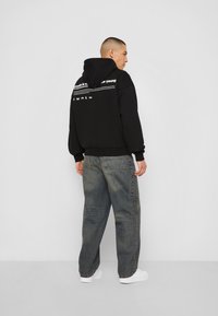 Black hoodie with front pocket, over-sized fit, featuring white text and graphic designs on the back, paired with loose light wash denim jeans.