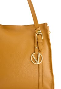 Tan leren schoudertas met een zachte textuur, verstelbare band en een gouden ronde logo-hanger. Heeft een minimalistisch ontwerp.