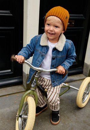 Ragazzo giovane con cappello di lana e giacca di jeans sorride mentre tiene il manubrio di una bici da equilibrio verde su un marciapiede cittadino.