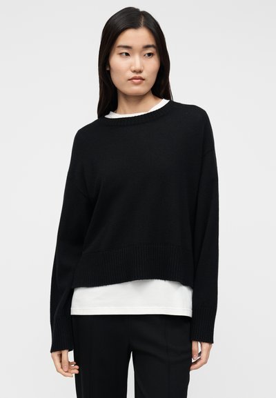 Pull noir en maille avec un large col bateau, ourlet raccourci et poignets côtelés. Porté sur un haut blanc. Texture lisse, coupe décontractée.