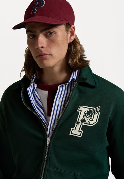 Giacca verde con zip e logo "P" ricamato e dettaglio a forma di ala, indossata sopra una camicia a righe. Cappello bordeaux con logo "P". Texture del tessuto morbida.