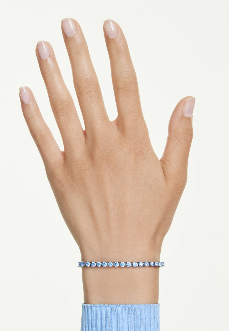 Main à peau claire portant un bracelet tennis serti de pierres précieuses bleues et une manche côtelée bleu clair sur un fond uni.