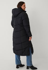 Zwarte pufferjas met een capuchon, met een gebreid ontwerp, lange lengte en zijsplitten. Gepaard met blauwe jeans en zwarte enkellaarsjes.