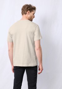 Světlé béžové tričko s krátkým rukávem vyrobené z bavlny, má kulatý výstřih a volný střih, v kombinaci s tmavými kalhotami, zobrazeno zezadu.