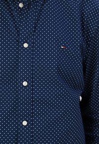 Navy blue button-up shirt with white polka dots. Features a small logo patch with red and white accents, and white buttons.