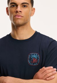 Navy modra bombažna majica s grafično printanimi palmami in besedilom v rdeči in svetlo modri barvi na levi strani prsnega področja.