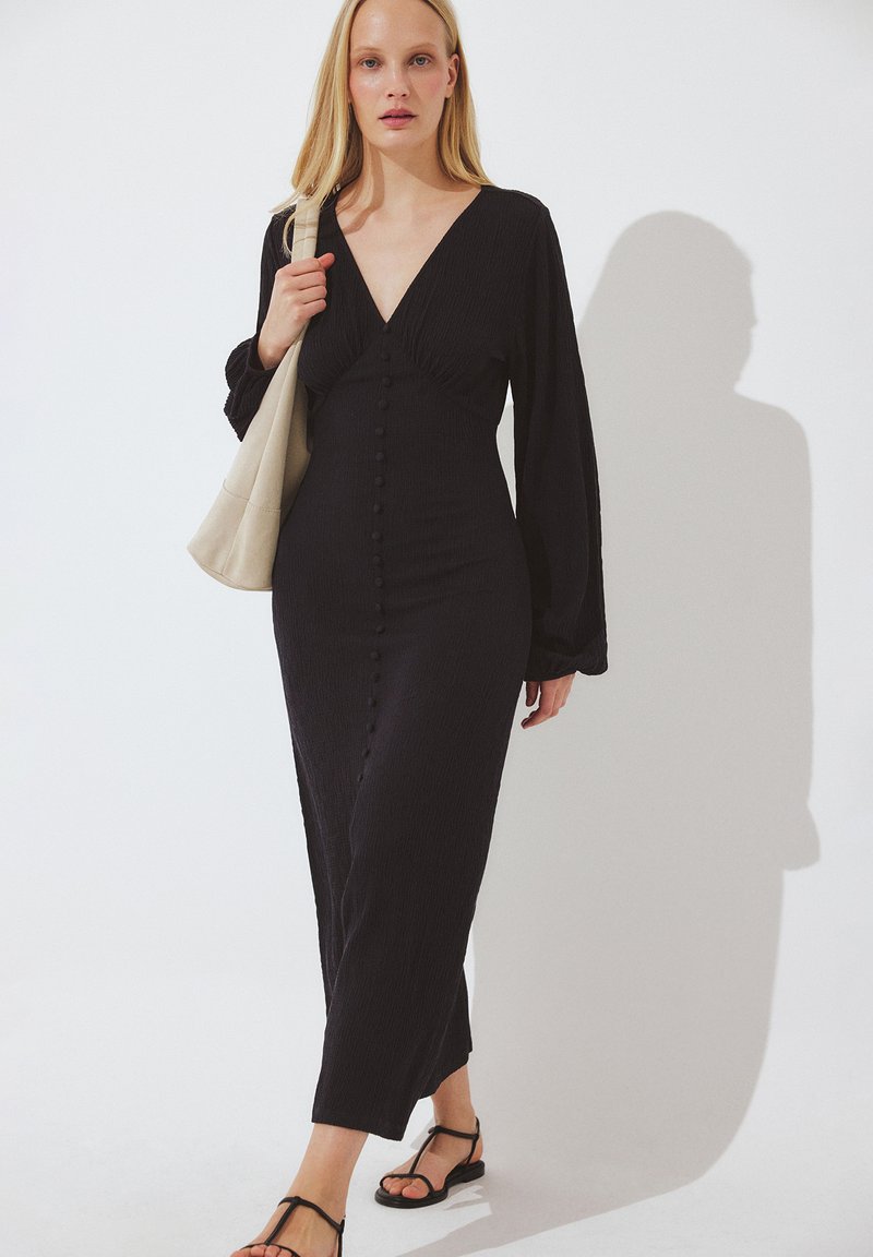 Naine, kes kannab pikemat musta nööbitavat kleiti ja musti sandaale, kandes beežikat õlakotti, kõnnib valge tausta ees.