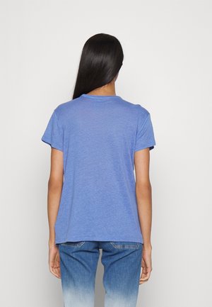 Persoon met lang donker haar staat met de rug naar ons toe, draagt een lichtblauw T-shirt met korte mouwen en tweekleurige blauwe spijkerbroek.