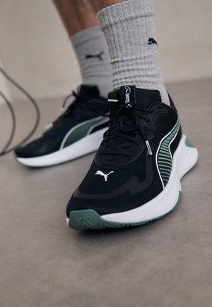 Osoba nosząca czarne i zielone buty sportowe Puma z białymi podeszwami oraz szare skarpetki z logo Puma na gładkiej podłodze.