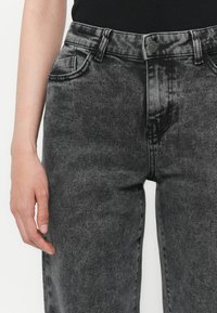 Close-up van een persoon die high-waisted, vervaagde zwarte jeans en een zwart topje draagt, met de linkertijd ontspannen naast hun lichaam.