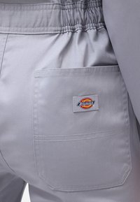 Zadní kapsa světle šedých kalhot s elastickým pasem, se čtvercovou nášivkou s logem značky Dickies v červené, žluté a černé barvě.