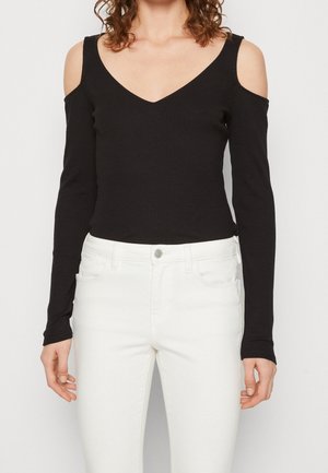 Frau trägt ein schwarzes Cold-Shoulder-Oberteil mit langen Ärmeln und V-Ausschnitt, kombiniert mit hoch taillierten weißen Jeans vor einem schlichten Hintergrund.