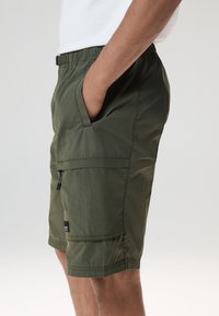 Pantaloncini verde oliva con tessuto leggero e testurizzato, vita elastica, tasche laterali e una piccola tasca con cerniera sulla coscia.