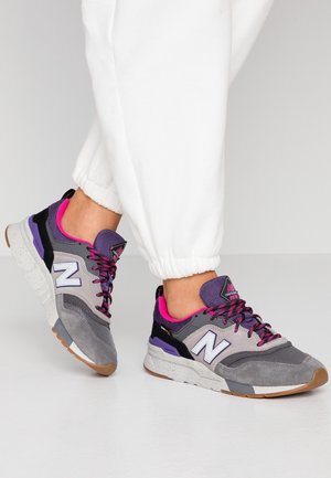 Jalat, joissa harmaat, violetit ja pinkit New Balance -lenkkarit, yhdistettynä valkoisiin collegehousuihin vaalealla taustalla.