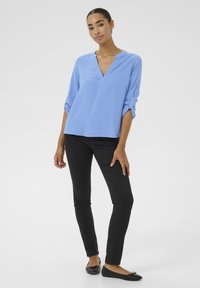 Blouse longue texturée bleu clair avec un décolleté en V, associée à un jean skinny noir et des ballerines noires. Les manches de la blouse sont retroussées.