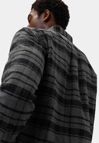 Grijze geruite blouse met zwarte accenten, gemaakt van structuurstof. Bevat een kraag en lange mouwen, met een close-up weergave van de schouder.