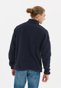 Tumman merensinisen fleece-takin, jossa on teksturoitu pinta, korkea kaulus ja pitkät hihat. Yhdistetty vaaleansinisiin farkkuihin, nähtynä takaa.