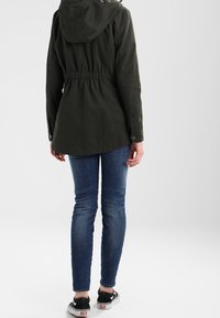 Personne portant une veste à capuche vert foncé, un jean skinny bleu et des baskets noires, debout de dos sur un fond clair uni.