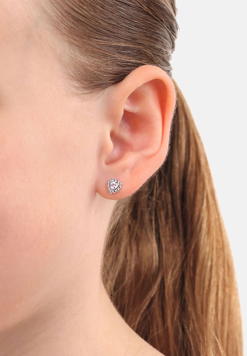 Närbild av en kvinnas öra med ljusbrunt hår som bär en liten hjärtformad silverörhänge med en rosa centrumsten.