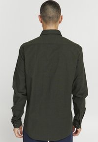 Camisa de manga larga verde oscuro con cuello, puños abotonados y corte recto. El material parece tener textura con un sutil brillo.