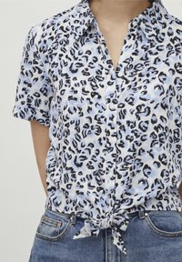 Donna che indossa una camicia a maniche corte con bottoni, stampata con macchie di leopardato blu e nero, annodata in vita, abbinata a jeans in denim blu.